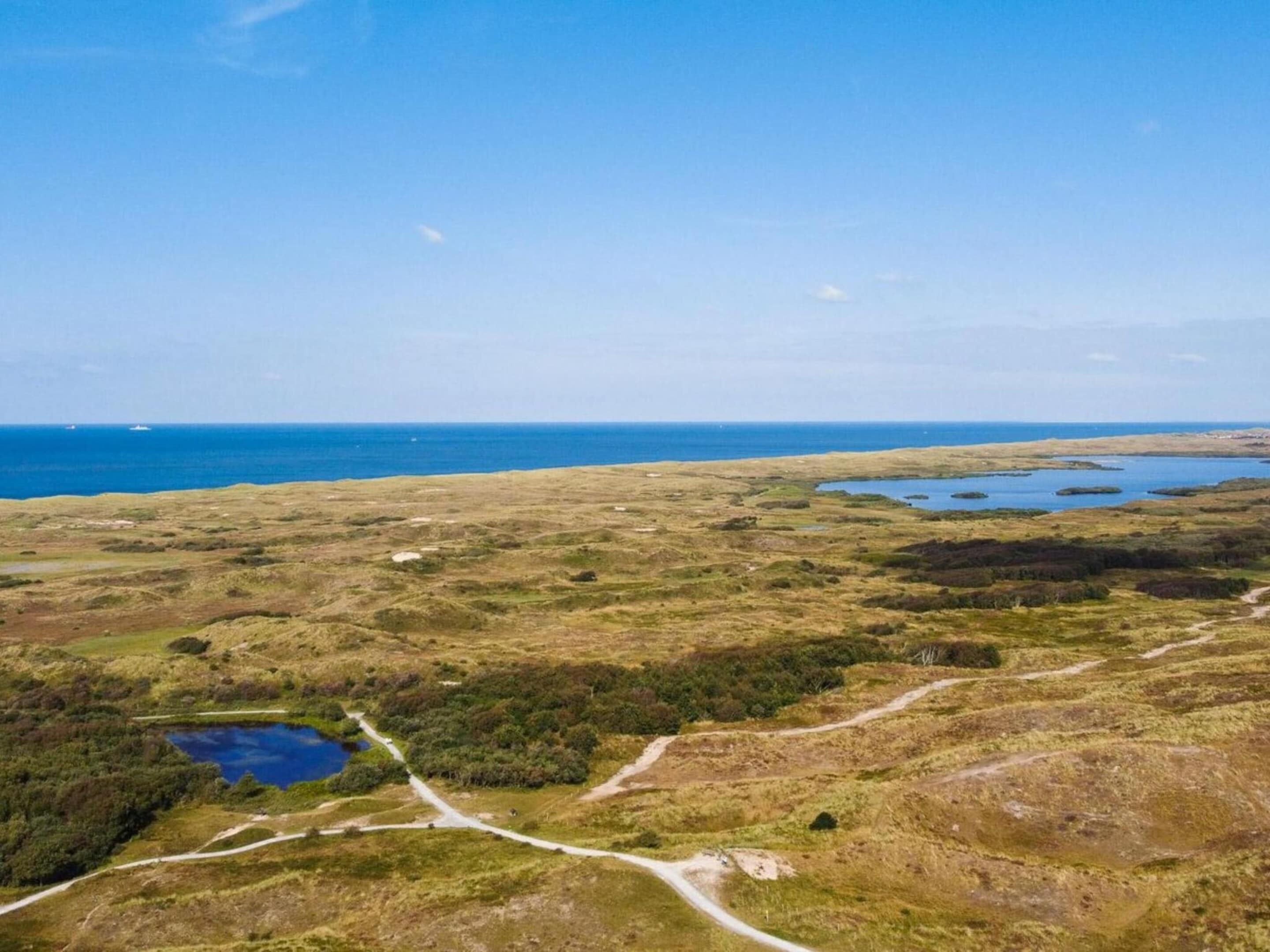An aerial view of a coastal landscape with a clear sky, a body of water, and a dirt path.