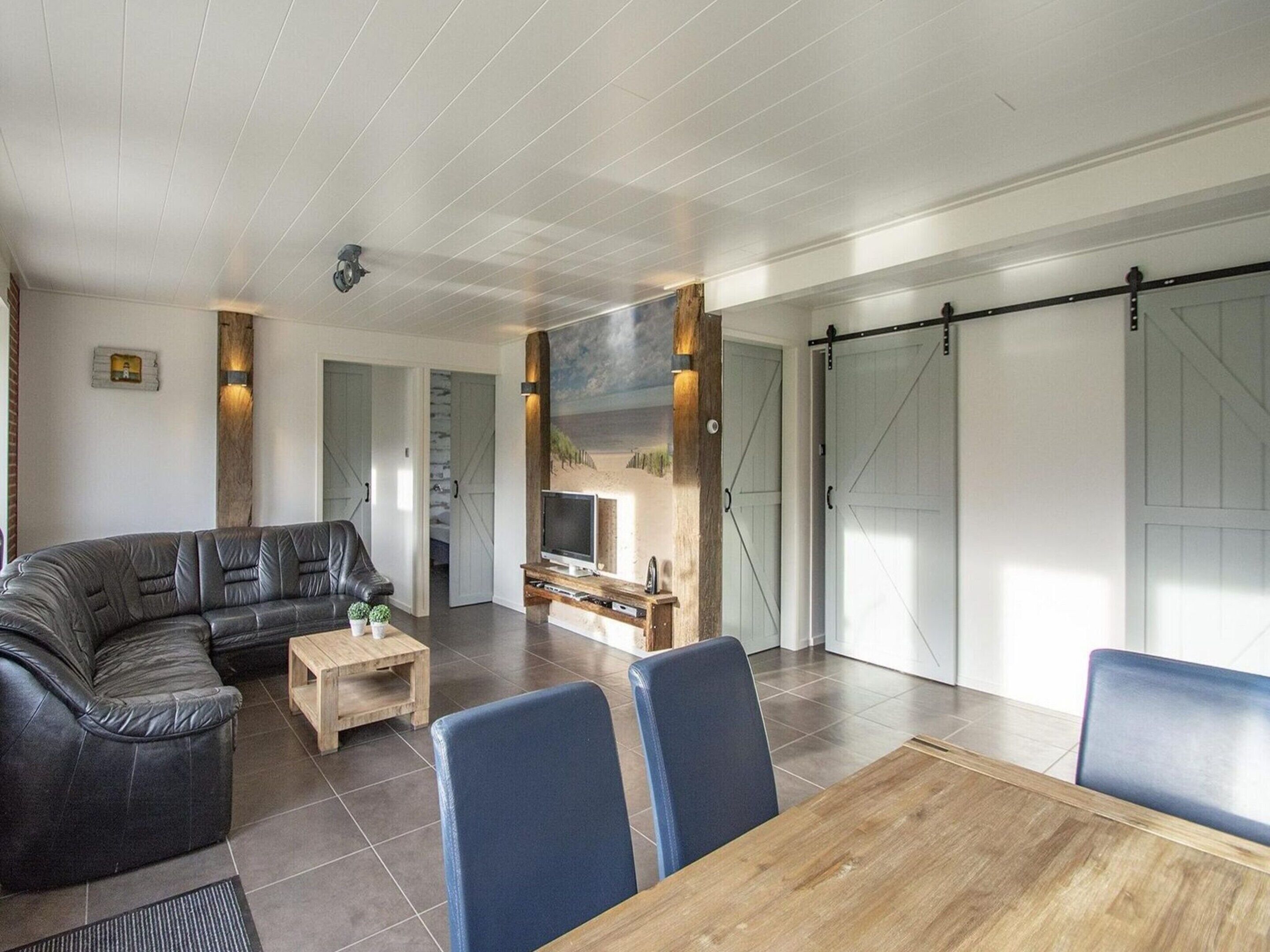 A modern living room with a leather sofa, a wooden coffee table, and a TV on a wooden stand. There are blue chairs around a wooden dining table.