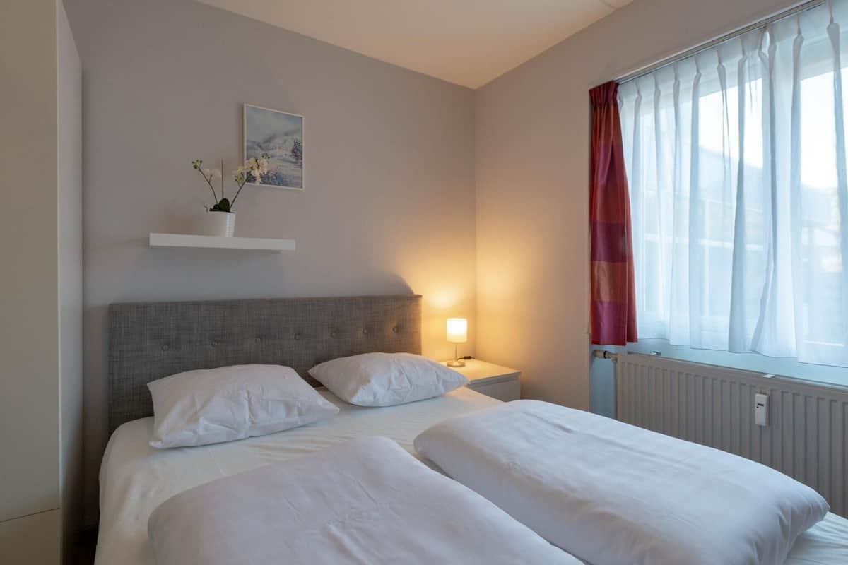 A hotel room with a double bed, a bedside table with a lamp, a small shelf with a plant, and a window with sheer curtains.