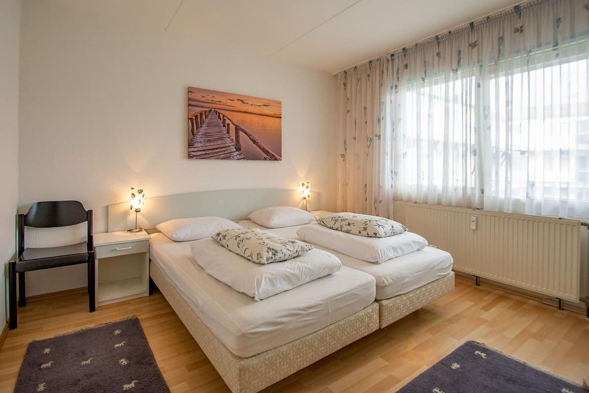 A bedroom with two beds, a chair, a nightstand, a lamp, a painting, and a window with curtains.