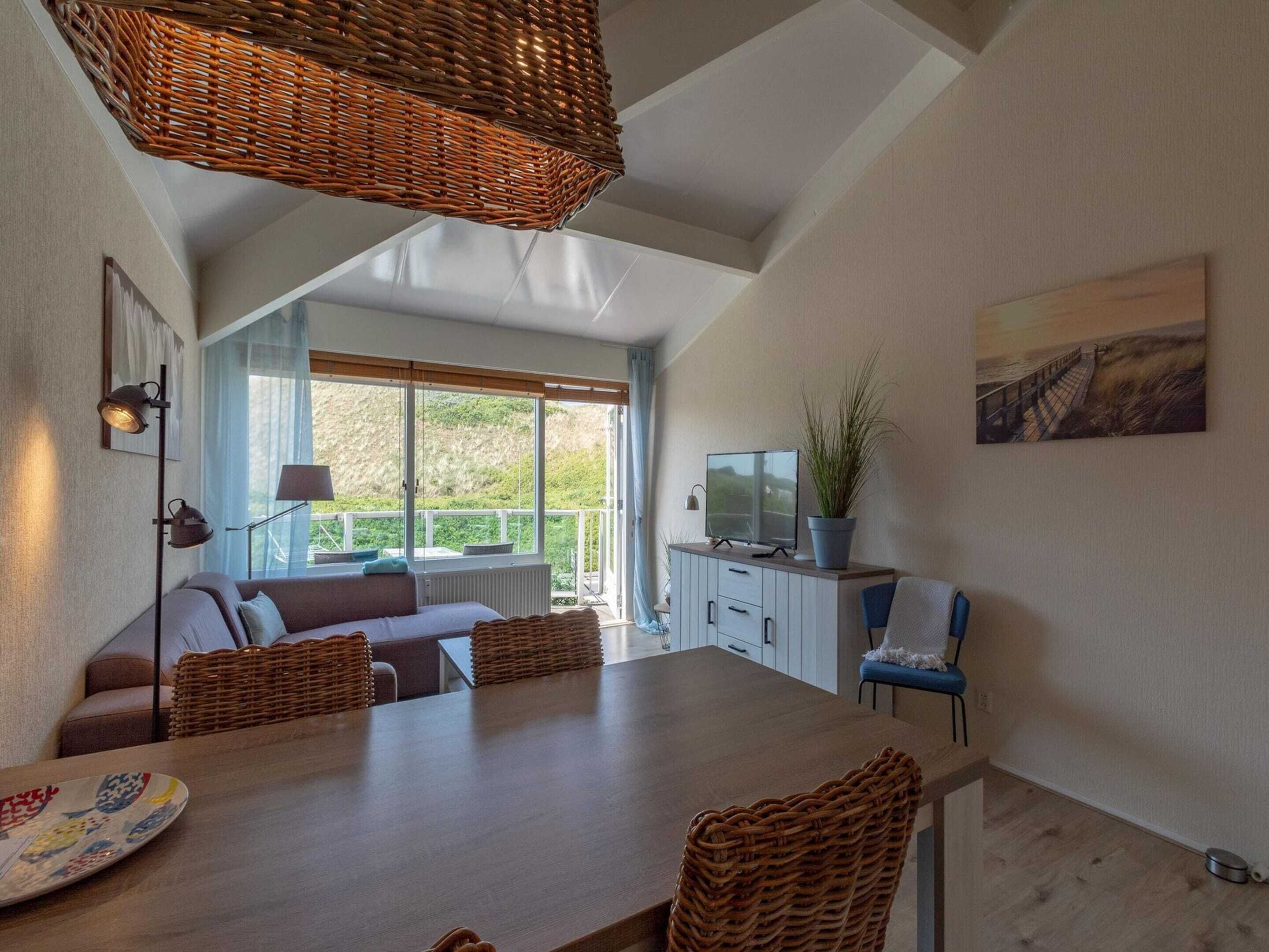 A modern living room with a large dining table, wicker furniture, a television, and a view of the outdoors.
