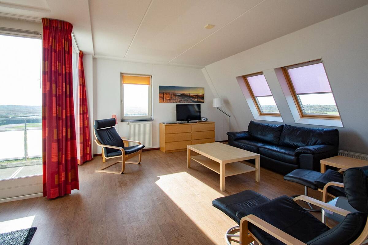 A modern living room with a sofa, chairs, a coffee table, and a TV. There are windows with curtains, a painting on the wall, and a view of the outdoors.
