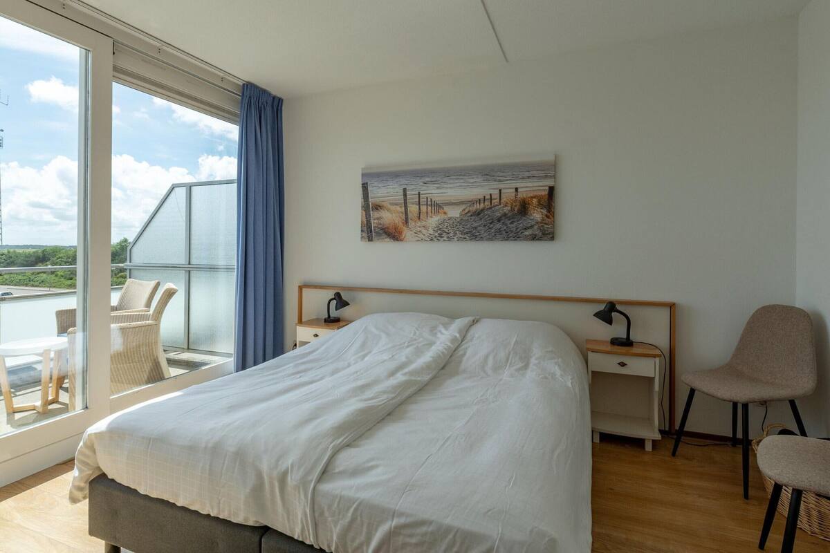 A hotel room with a large bed, a desk, a chair, and a balcony with a view of the sea.
