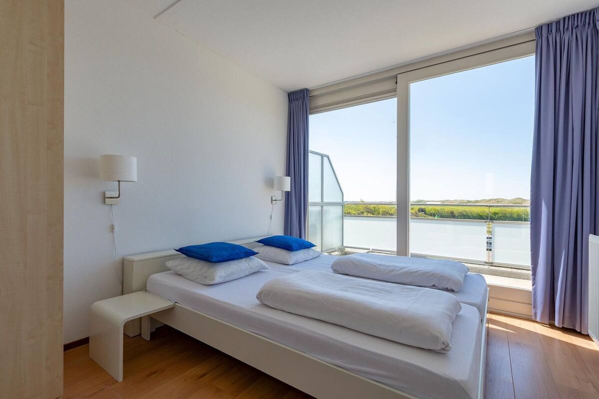 A modern bedroom with a large bed, two bedside tables, a bench, and a view of a body of water through large windows.