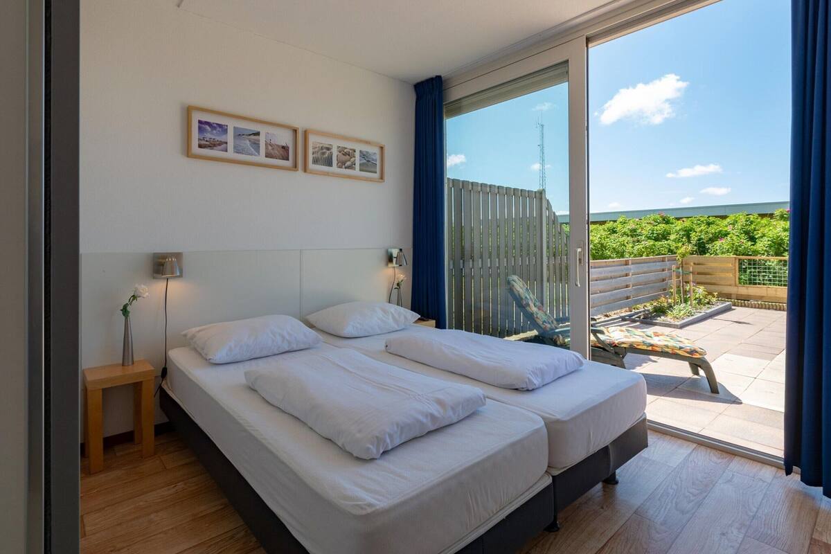 A bedroom with two beds, a small bedside table, a wall-mounted lamp, and a large sliding glass door leading to an outdoor area with a wooden deck and greenery.