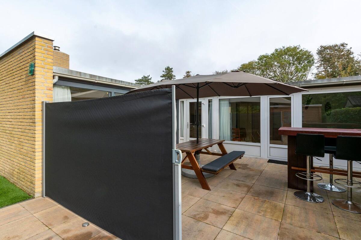 Un patio con mesa y bancos de madera, una zona de estar cubierta y una puerta de cristal que da a un espacio interior.