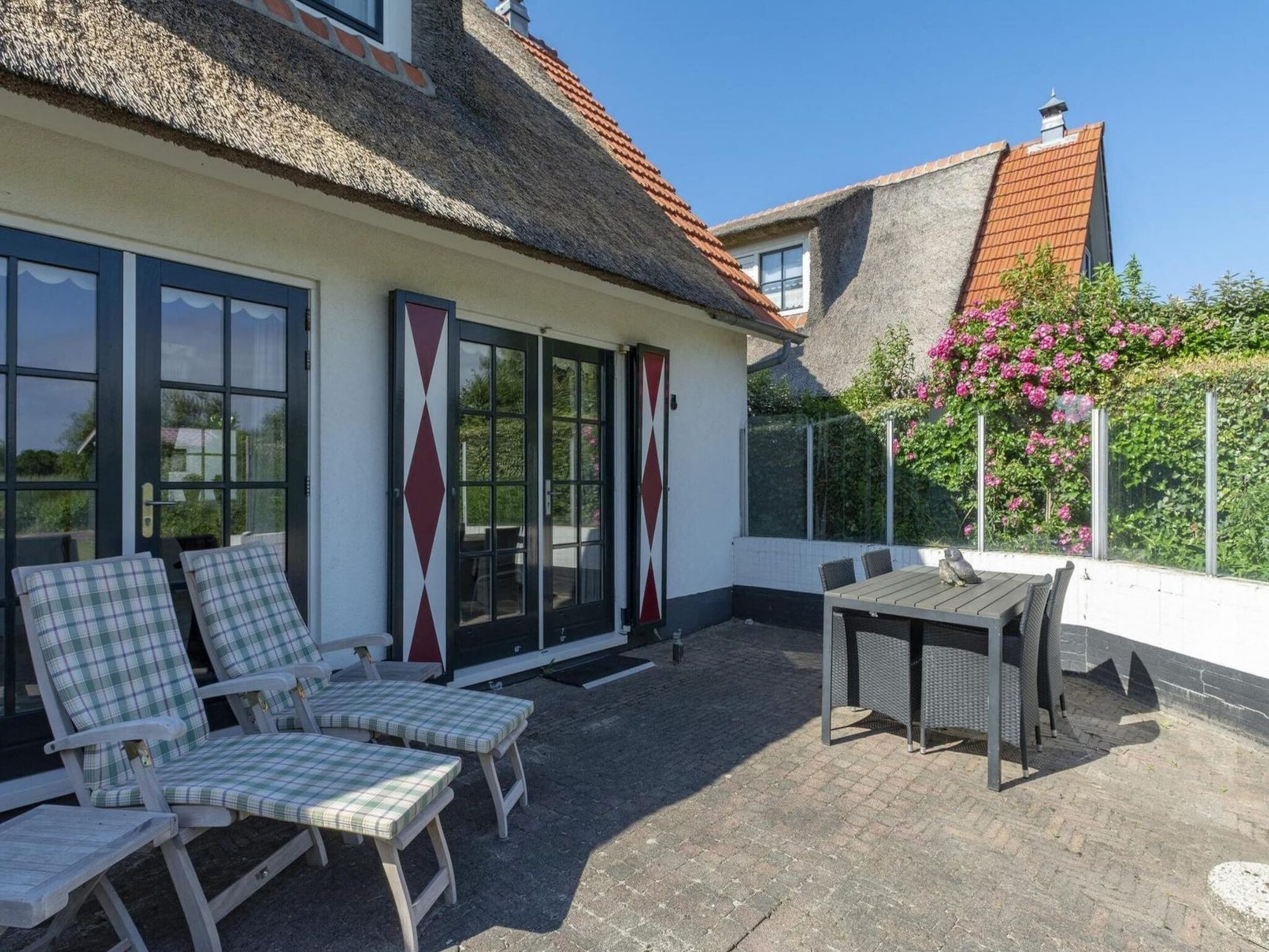 A patio with lounge chairs and a table, a thatched roof, and a garden with pink flowers.