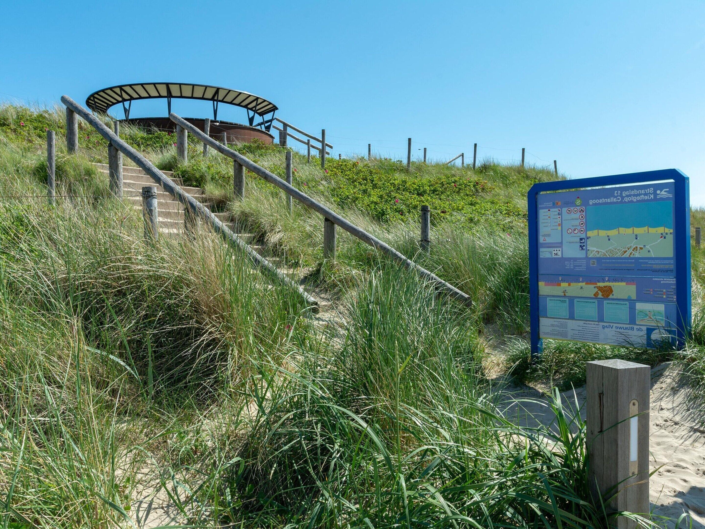 Eine Treppe am Strand, die zu einem überdachten Bereich führt, in dessen Nähe ein Wegweiser steht.