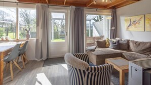 Ein gemütliches Wohnzimmer mit Holzbalkendecke, einer Couch, einem gestreiften Sessel, einem Esstisch mit Stühlen und einem großen Fenster mit Vorhängen.
