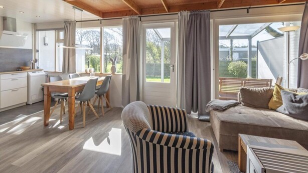Eine moderne Küche mit Holzbalken, einem Essbereich mit Tisch und Stühlen, einem gestreiften Ohrensessel, einer Couch mit Kissen und einem großen Fenster mit Vorhängen.