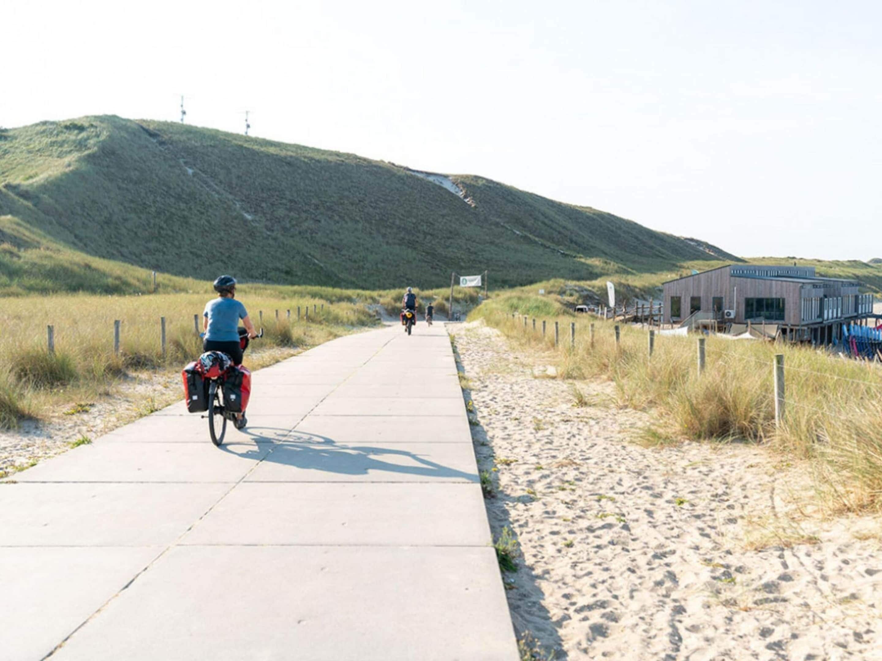 A paved pathway alongside a sandy beach with a hill in the background.