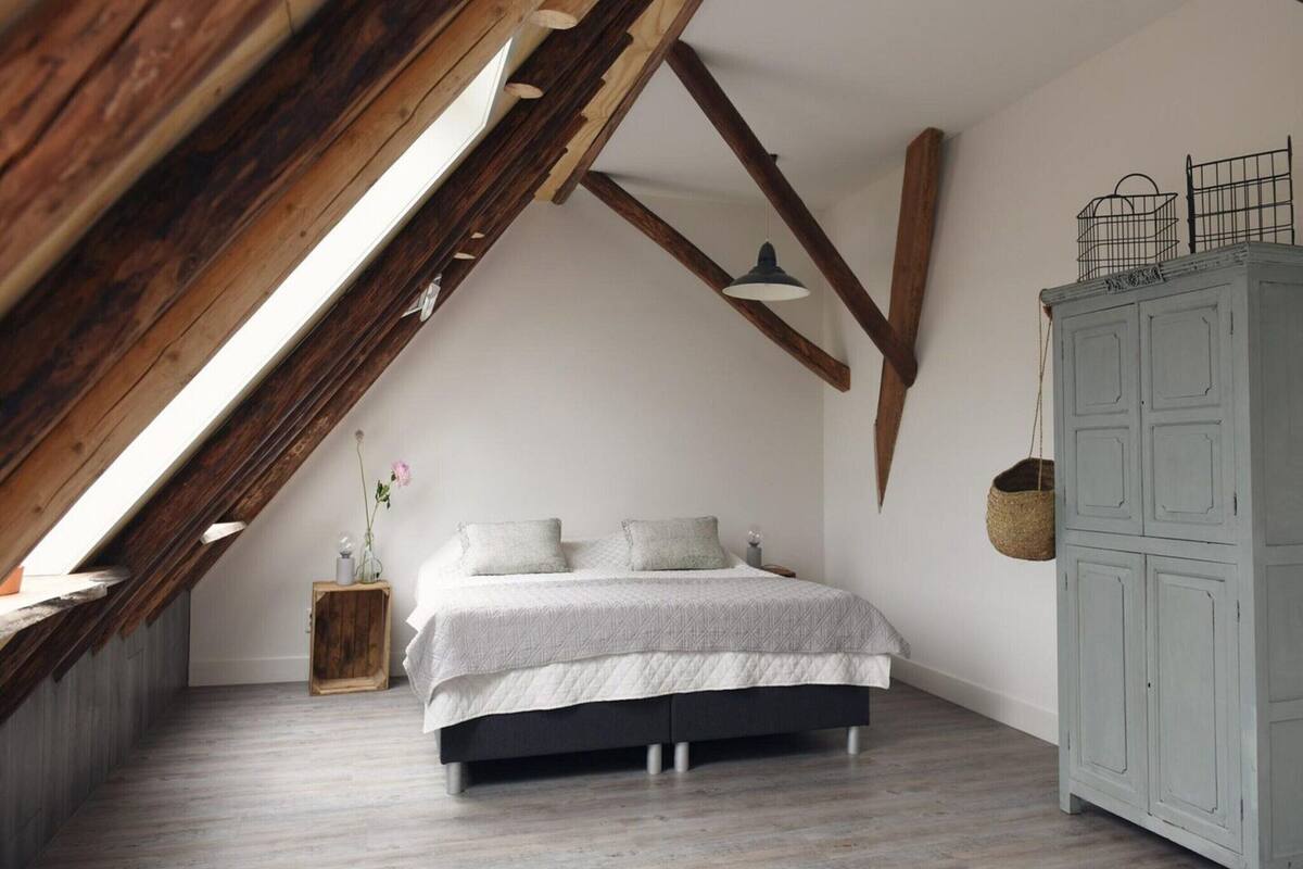 Ein Schlafzimmer mit schrägem Dach, einem Bett mit weißer Bettdecke, einem grauen Kleiderschrank und einem kleinen Nachttisch aus Holz.