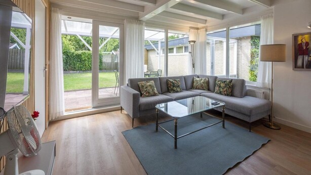 A spacious living room with a grey sofa, a glass coffee table, and a large sliding glass door leading to a garden. - Comfortable Holiday Home in Sint Maartenszee (Sint Maartensvlotbrug)