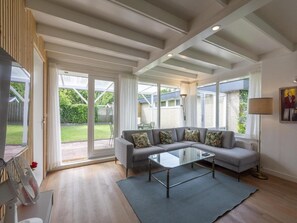 A spacious living room with a grey sofa, a glass coffee table, and a large sliding glass door leading to a garden. - Comfortable Holiday Home in Sint Maartenszee (Sint Maartensvlotbrug)