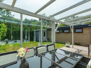 A conservatory with a glass table and chairs, a vase with flowers, and a view of a garden and a brick wall.