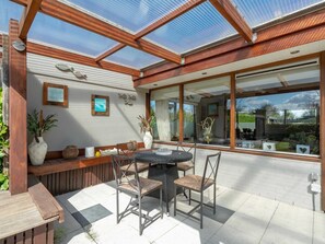 A sunlit patio with a wooden table and chairs, a bench, and a glass-enclosed area with a view of the outdoors.