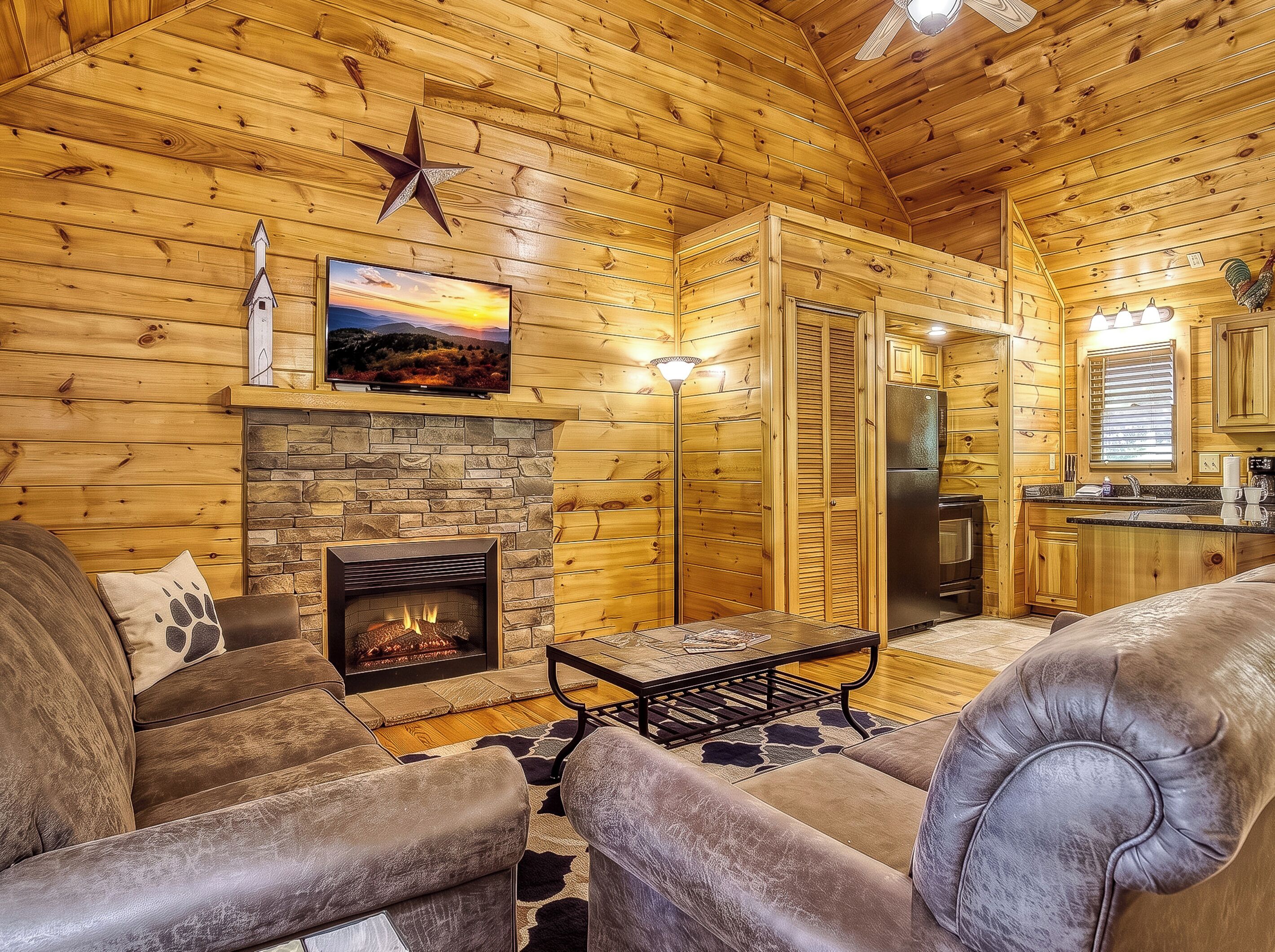 A log cabin interior with a stone fireplace, leather furniture, a flat-screen TV, and a kitchen area.