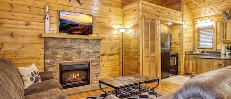 A log cabin interior with a stone fireplace, leather furniture, a flat-screen TV, and a kitchen area.