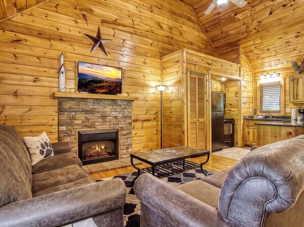 A log cabin interior with a stone fireplace, leather furniture, a flat-screen TV, and a kitchen area.