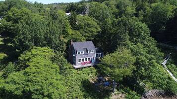 A two-story house surrounded by dense forest, with a visible deck and outdoor seating area.