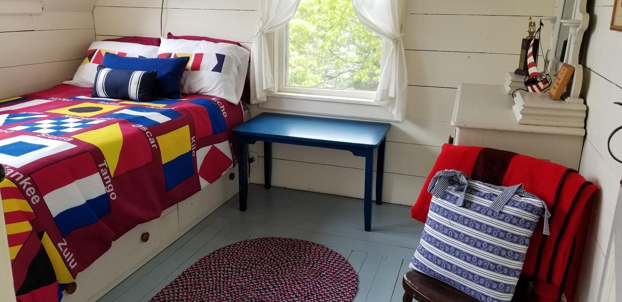 Een kamer met een bed voorzien van een kleurrijke sprei, een kleine tafel, een stoel met een rode kussen en een raam met gordijnen.