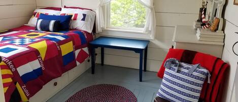 Een kamer met een bed voorzien van een kleurrijke sprei, een kleine tafel, een stoel met een rode kussen en een raam met gordijnen.
