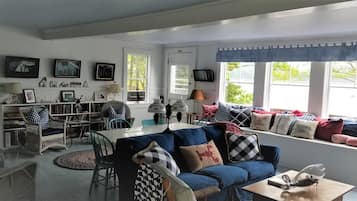 A cozy living room with a blue sofa, wicker chairs, a dining table, and a window with curtains.