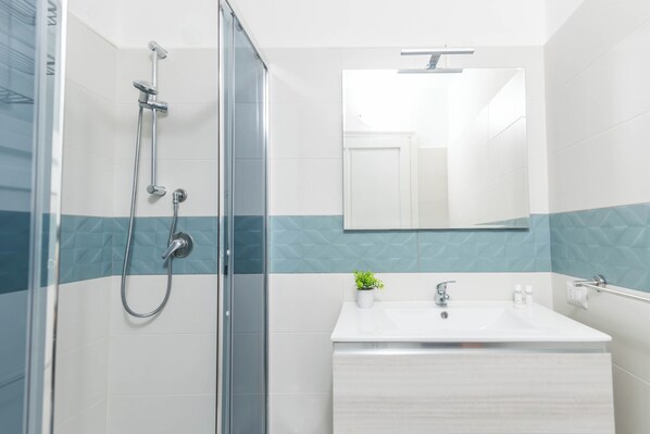 Une salle de bain moderne avec une douche à l’italienne en verre, un lavabo blanc avec un miroir au-dessus, et une petite plante en pot posée sur le lavabo.