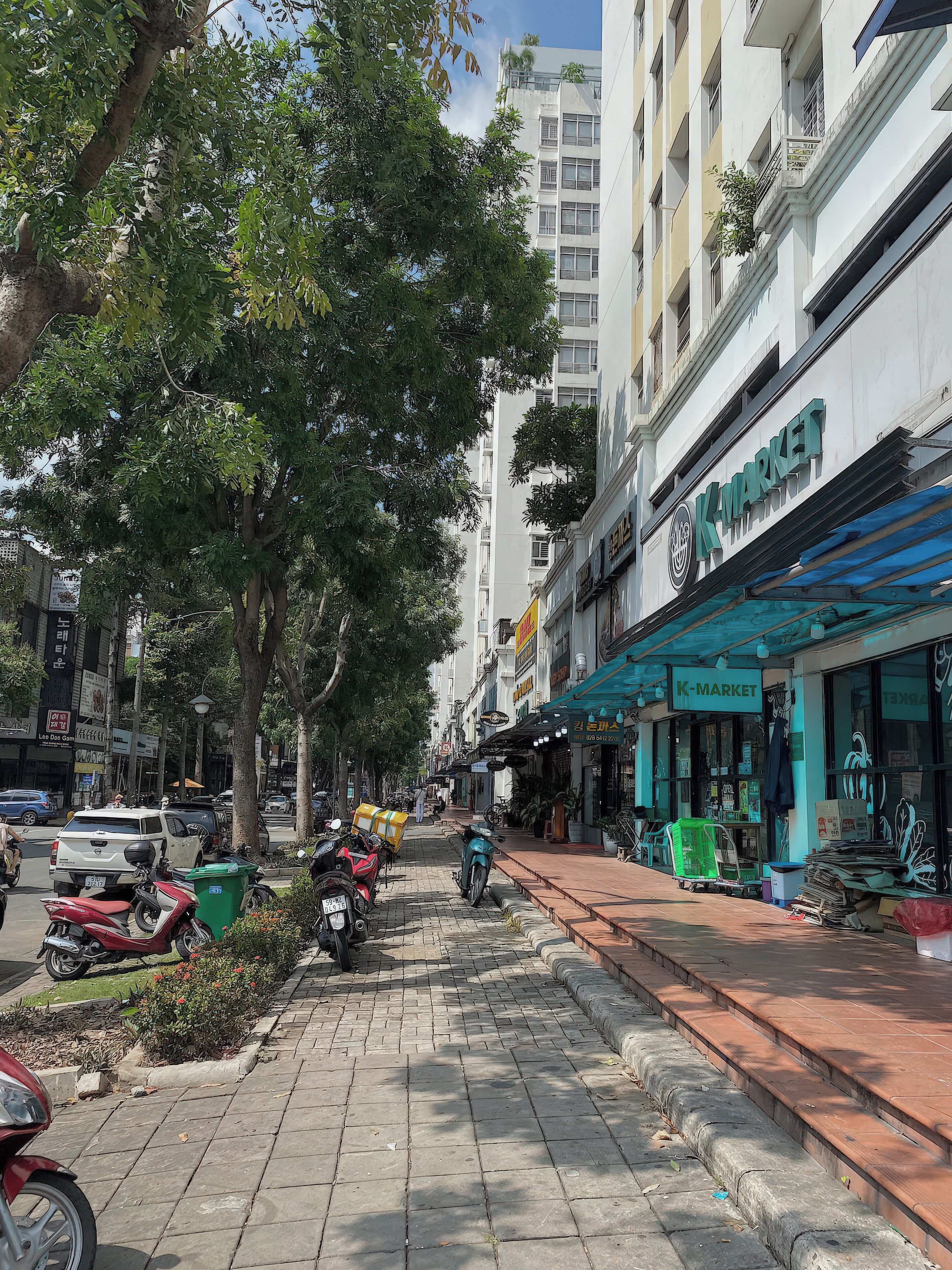 A street with a K-Market store, parked motorcycles, and a sidewalk lined with trees.