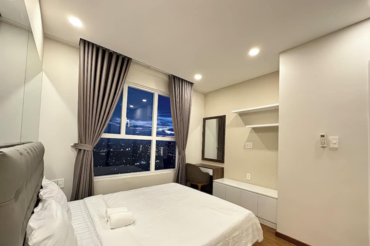 Un dormitorio moderno con una cama grande, un escritorio, una silla, una mesita de noche y un ventanal con cortinas.