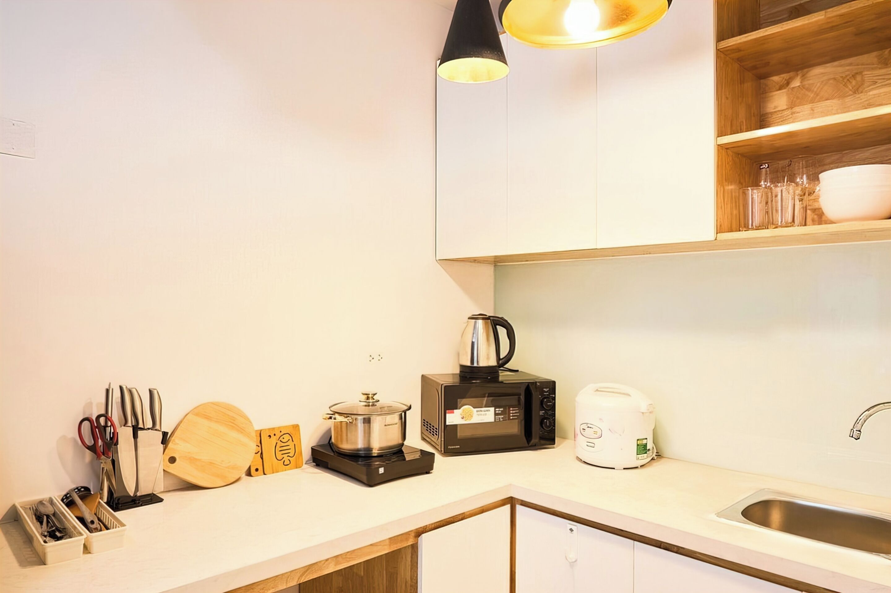 Una cocina con microondas, tostadora y hervidor sobre la encimera, un lavabo y un estante de madera encima.