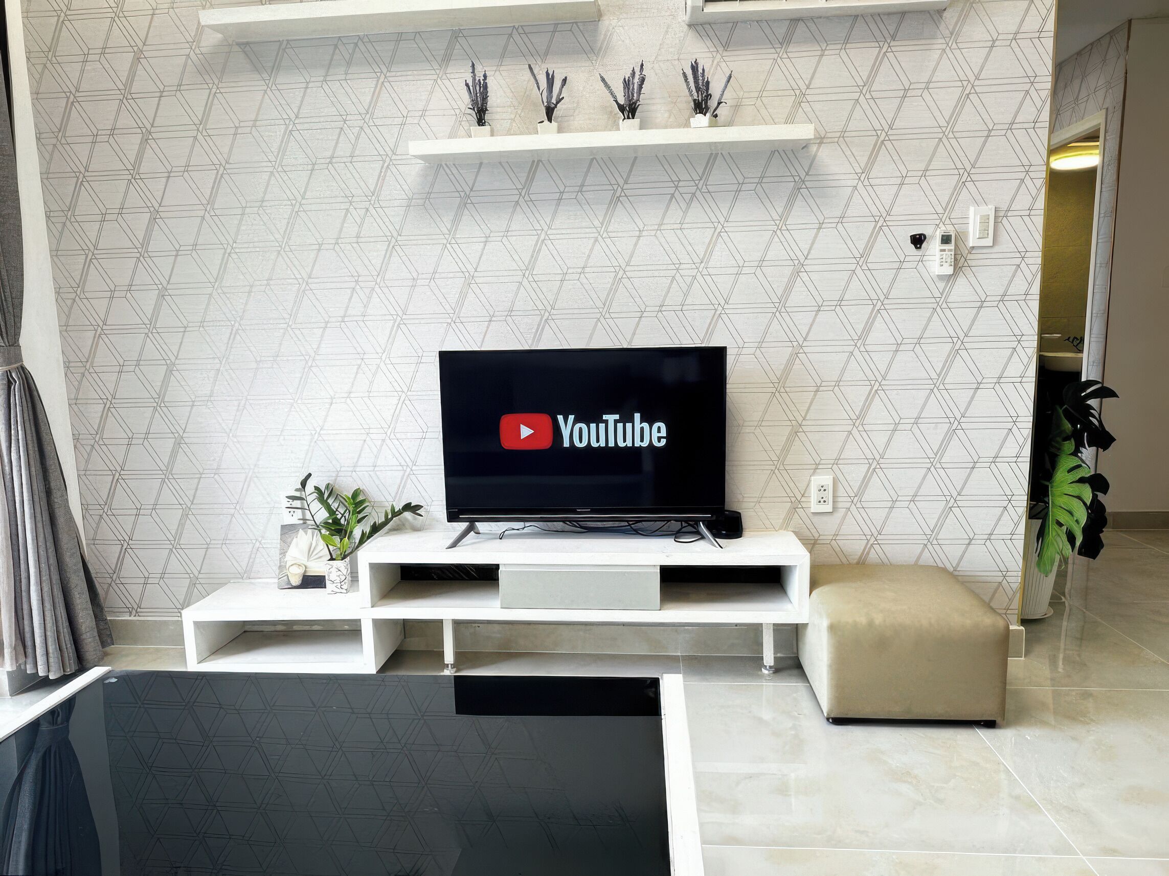 Moderni olohuone, jossa on litteänäyttöinen televisio, jonka ruudulla näkyy YouTuben logo, valkoinen mediakeskus, beigenvärinen jalkarahi ja geometrisesti kuvioitu seinä.