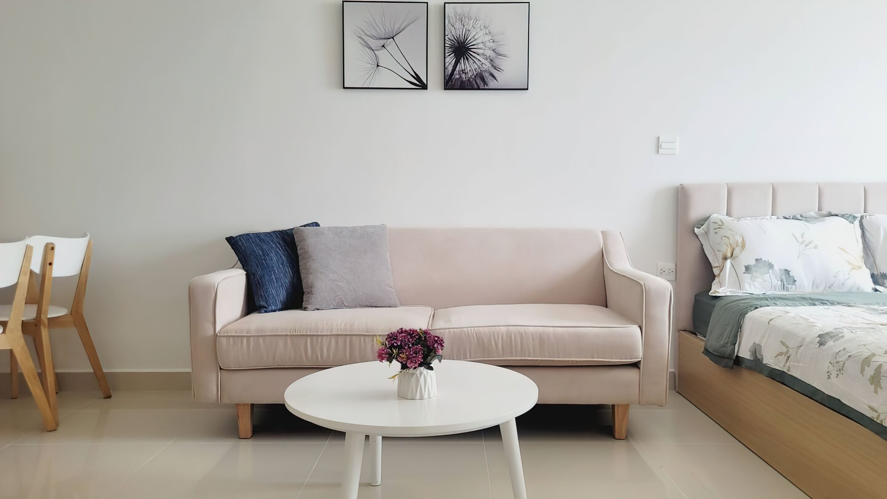 A living room with a beige sofa, a round white coffee table, and a bed with floral bedding. There are two framed pictures on the wall and a vase with flowers on the coffee table.