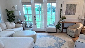 Una sala de estar con un amplio sofá modular, un reposapiés redondo, una mesita pequeña y una silla. Se aprecia una ventana grande con vistas a un jardín.