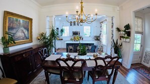 Comedor con una mesa grande, sillas y un candelabro. Se ve una cocina al fondo con armarios y una ventana.