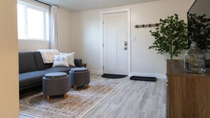 Un salon avec un canapé gris, deux poufs, un tapis à motifs, un meuble en bois orné d’une plante et une porte blanche.