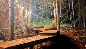 A wooden deck with benches in a forested area, surrounded by trees and illuminated by string lights. - Little Creek Cabin: Cozy, Adventure-Ready & Dog-Friendly (Anchorage)