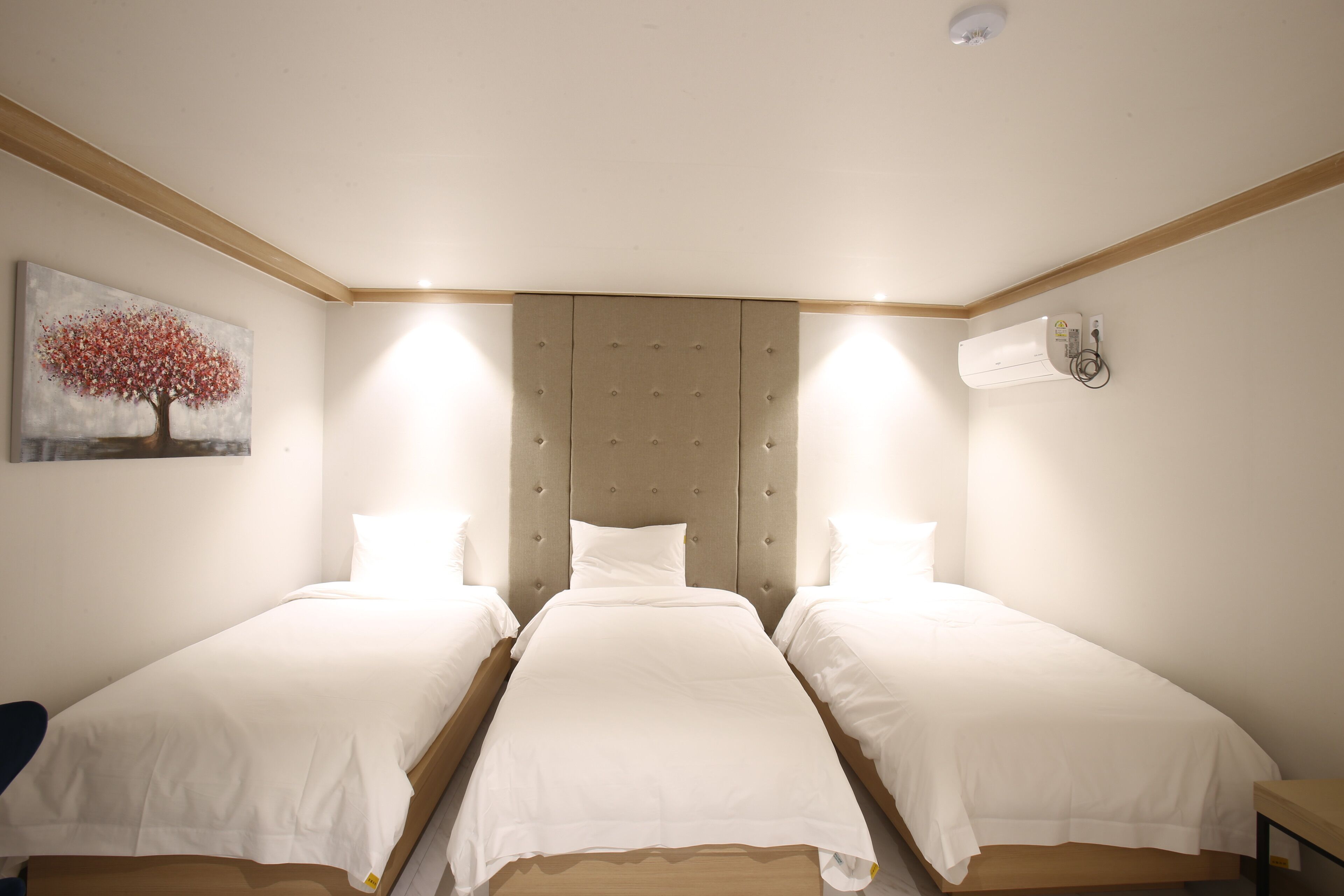 A room with three single beds, a headboard, a painting of a tree, and a wall-mounted air conditioning unit.