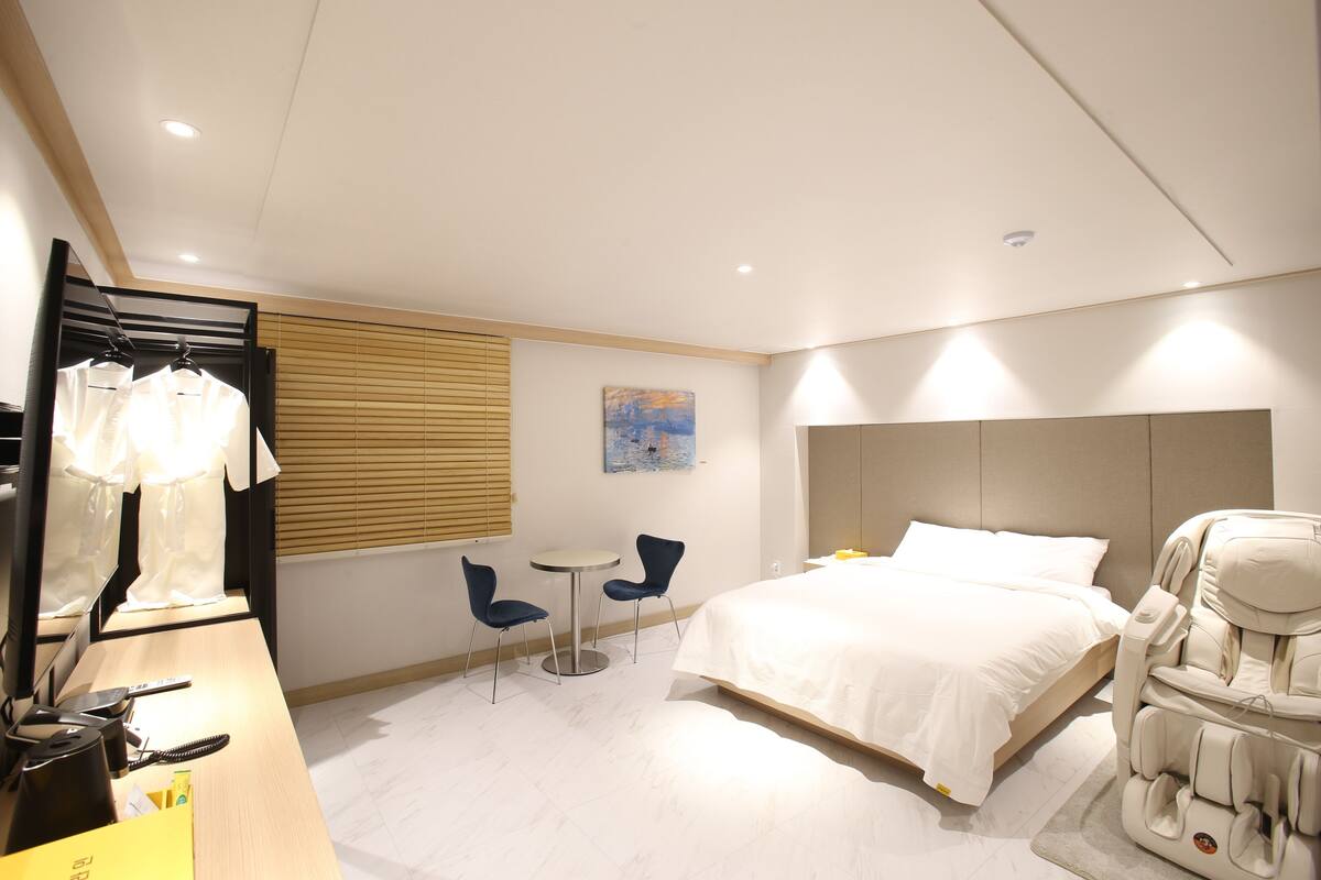 Ein modernes Hotelzimmer mit einem Bett, Massagesessel, zwei Stühlen, einem kleinen Tisch und einem Spiegel.