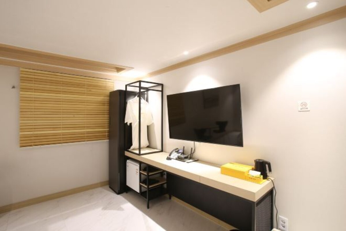 A modern bedroom with a bed, a desk, a chair, a TV, and a window with blinds.