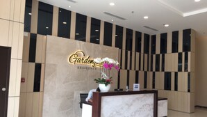 Un comptoir d’accueil surmonté d’un plateau en marbre et agrémenté d’une orchidée en pot. Derrière le comptoir, un mur est décoré de panneaux verticaux noirs et beiges, et une enseigne indique : « Gardenzoo Residence ».