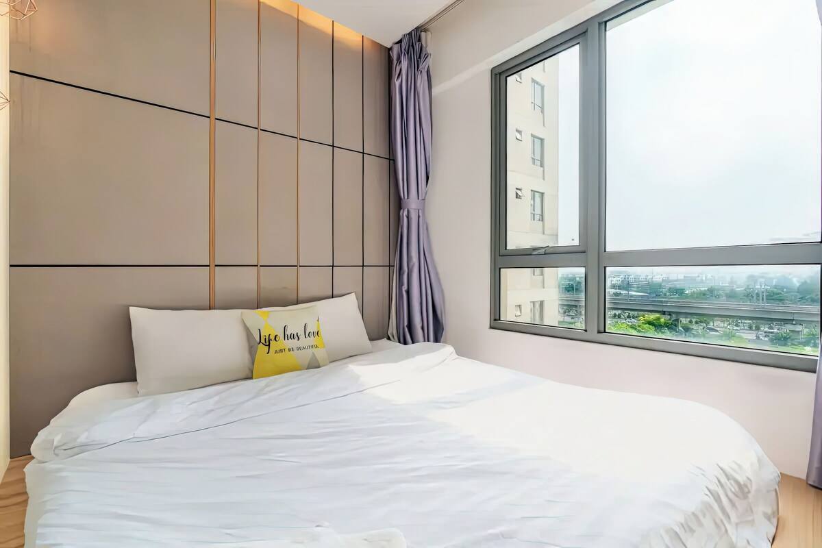 Une chambre moderne avec un grand lit, une vue sur la ville par la fenêtre et un panorama urbain visible à l’extérieur.