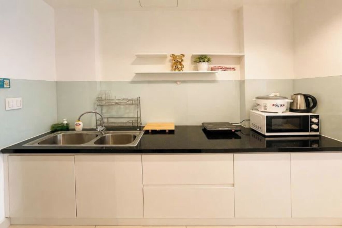 A kitchen with a black countertop, stainless steel sink, white cabinets, a microwave, and a kettle.