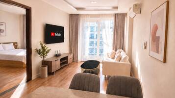 Moderni hotellihuone, jossa on sänky, seinälle asennettu televisio, ruokailutila pöydän ja tuolien kera sekä näkymä kaupunkiin suurista ikkunoista.