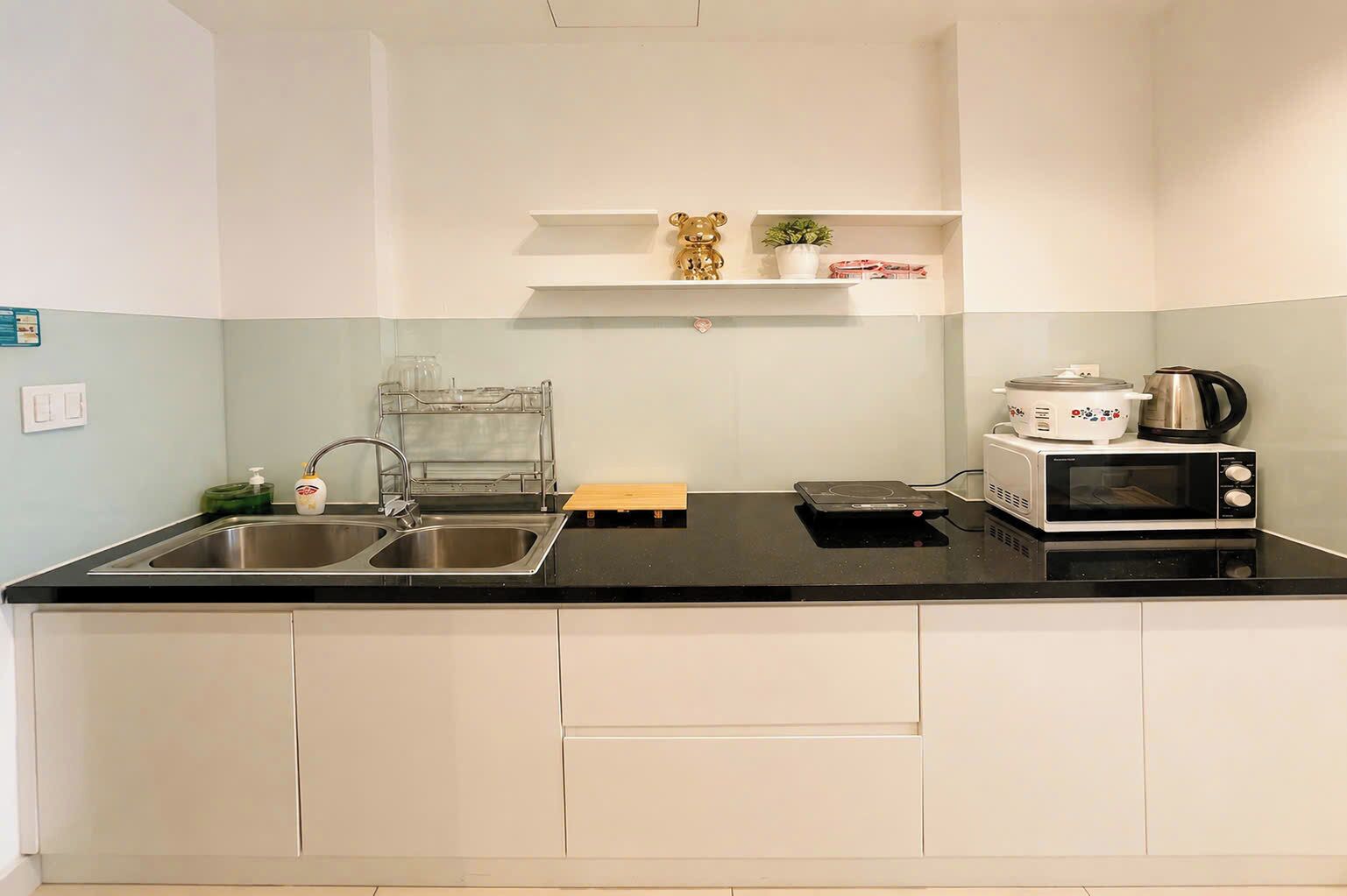 A kitchen with a double sink, a microwave, a kettle, and a small shelf with decorative items.
