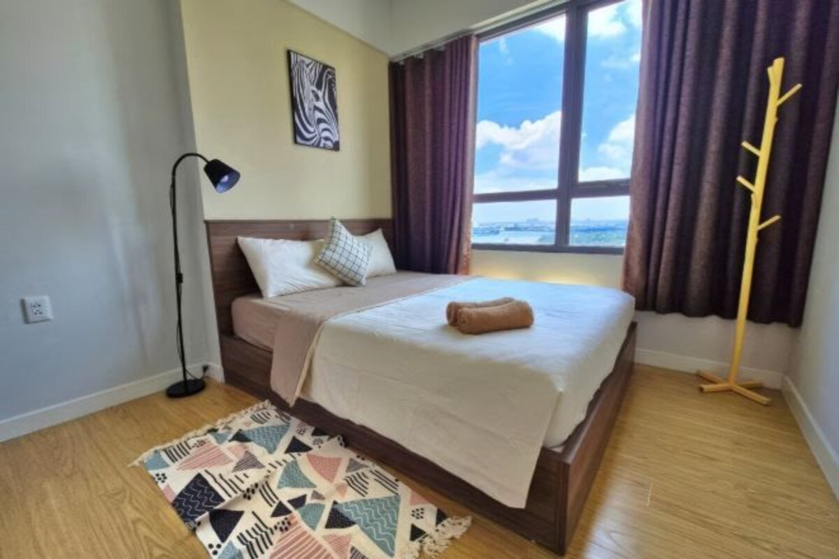 A bedroom with a bed, a floor lamp, a window with curtains, and a view of the sky.