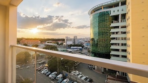 Um edifício alto com fachada de vidro curva, um estacionamento com carros e um horizonte urbano com edifícios e árvores ao fundo.