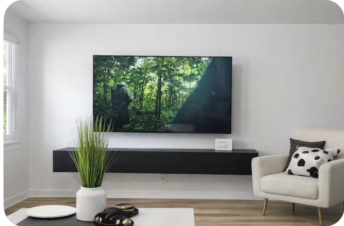 A living room with a flat-screen TV mounted on a wall, a white armchair with a black and white patterned pillow, a small white table with a plant and a plate, and a black shelf below the TV.
