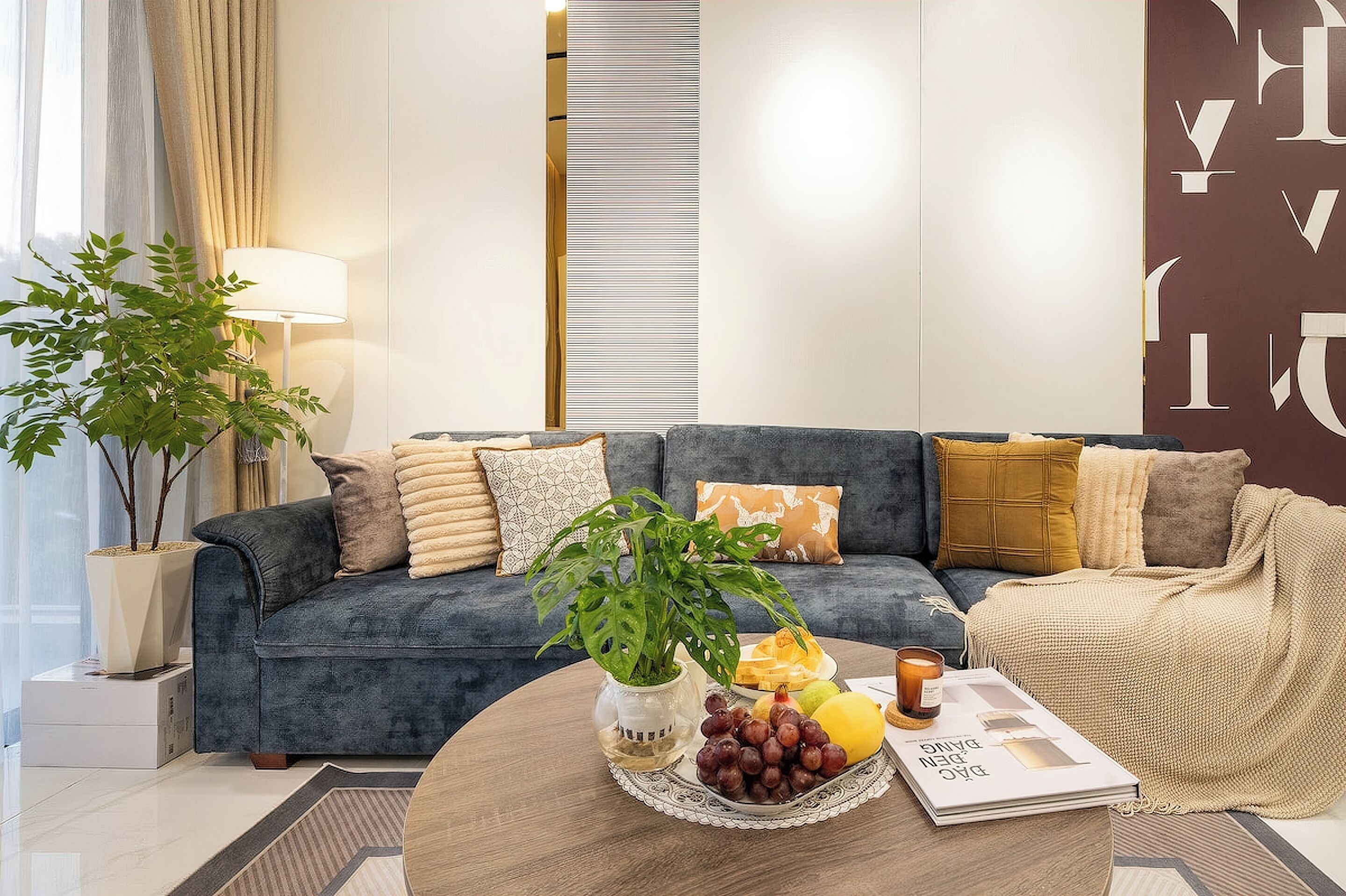 A modern living room with a dark blue sofa, decorative pillows, a potted plant, a fruit bowl, and a magazine on a coffee table.