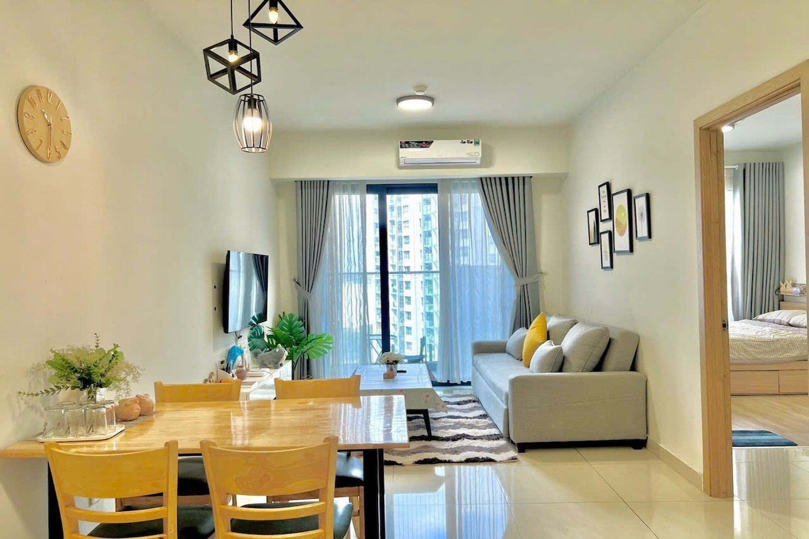 Una sala de estar moderna con un sofá, una mesa de comedor y sillas. Hay un ventanal con cortinas, un equipo de aire acondicionado y un reloj en la pared.